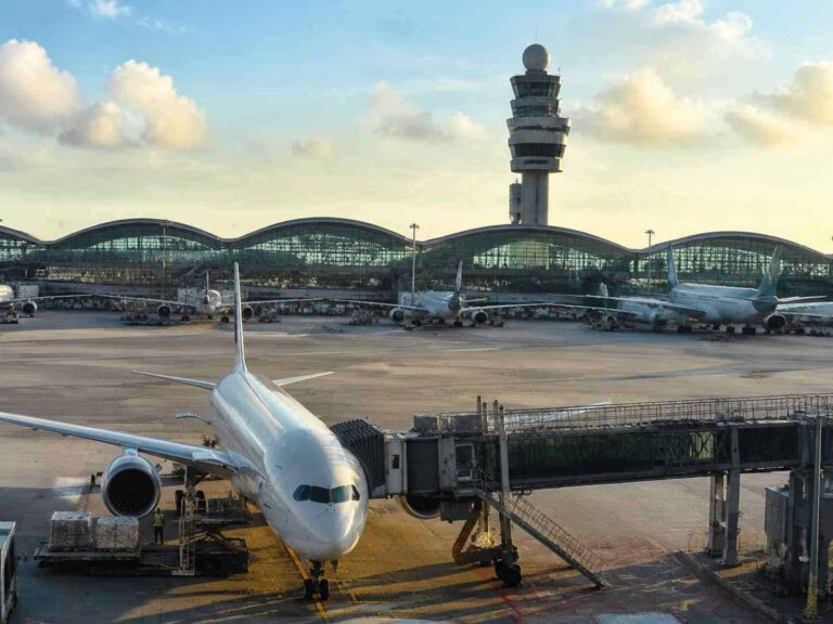 A large passenger jet is docked at a gate with a jet bridge connected, others parked along the tarmac and a control tower in the background at a sunny airport.