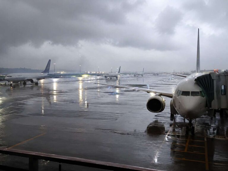 Airplane at a wet tarmac under dark cloudy skies, jet bridge connected to the foreground aircraft, distant planes parked along the runway
