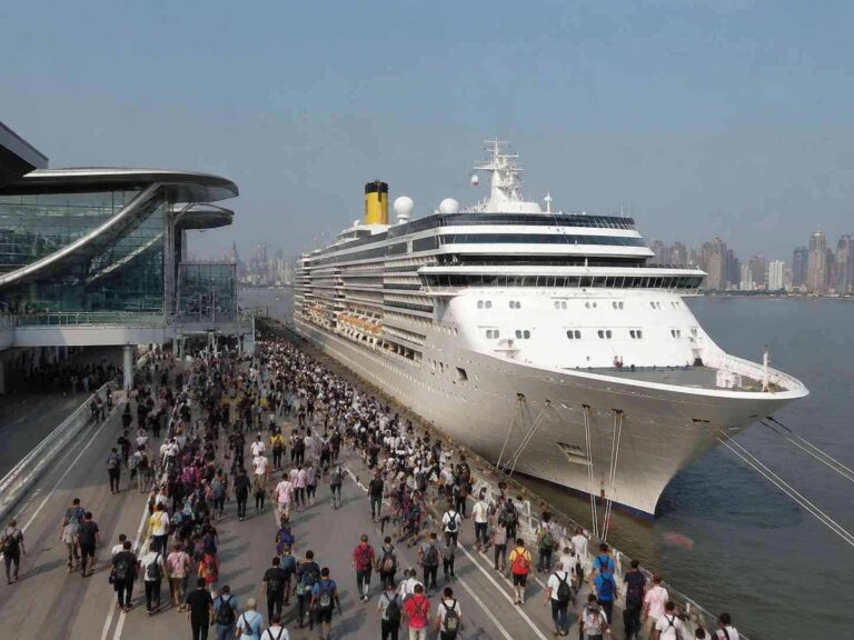 Large cruise ship docked at a busy port with a crowd walking along the pier and city skyline in the background
