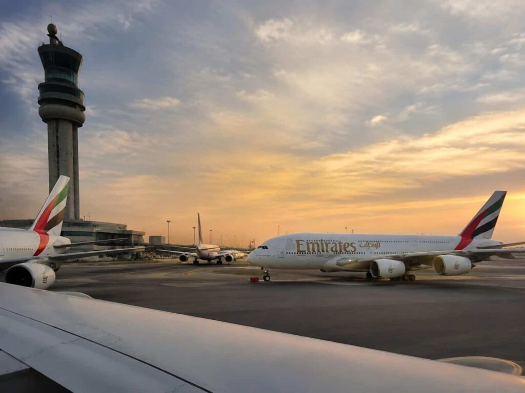 Emirates aircraft parked on a sunlit airport tarmac with a tall control tower in the background at sunset.