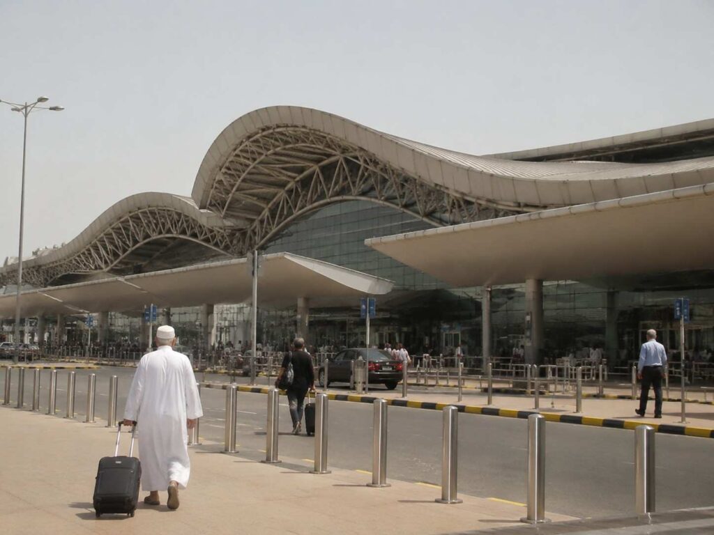 Traveler in white traditional clothing pulling a suitcase toward a modern airport terminal with a wavy roof of metal and glass.