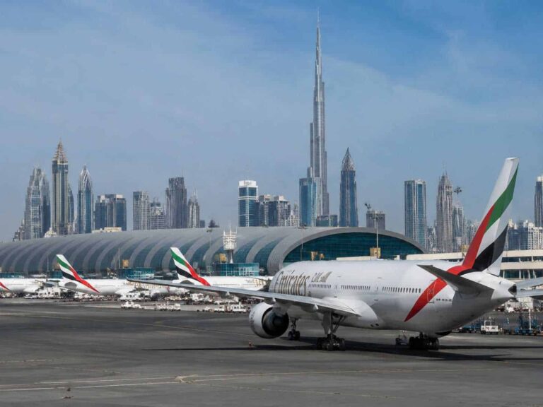 Wide view of Dubai International Airport with multiple Emirates planes on the tarmac and the futuristic city skyline, including the Burj Khalifa, in the background