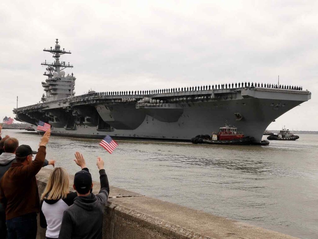 A gray US Navy aircraft carrier glides by as people on the shore wave American flags in celebration.
