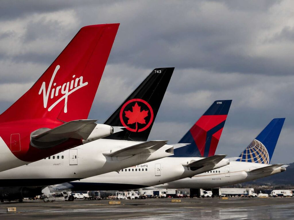 Tail fins of three commercial jets parked at an airport, featuring Virgin red tail, black tail with red maple emblem, and a blue globe-marked tail