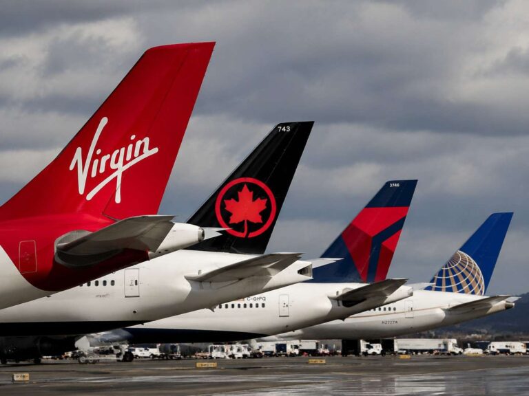Tail fins of three commercial jets parked at an airport, featuring Virgin red tail, black tail with red maple emblem, and a blue globe-marked tail