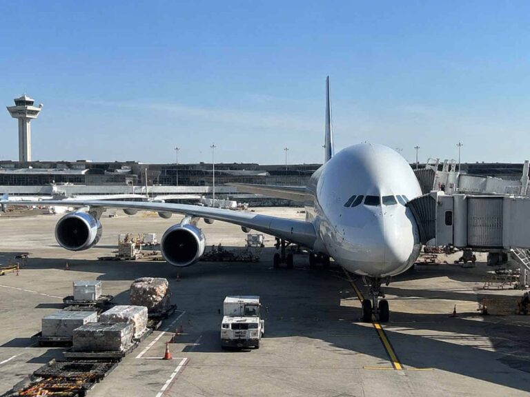 Four-engine wide-body airliner parked at a terminal gate with a jet bridge connected on the right and a control tower in the distance.