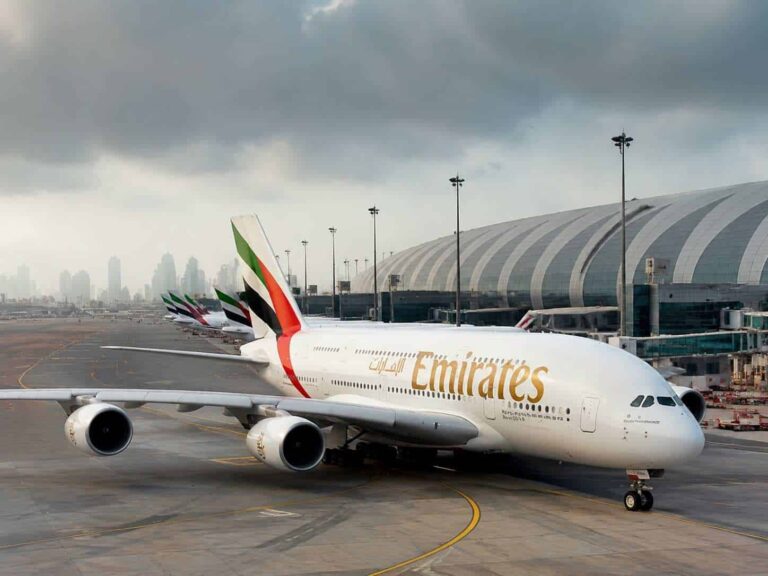 Emirates airliner parked at the gate on a cloudy day, with multiple aircraft tails in UAE colors lined up behind it.