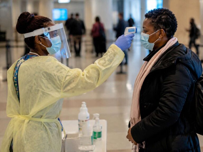 Healthcare worker in PPE uses a forehead thermometer to check a masked woman's temperature at a screening table in a public space.