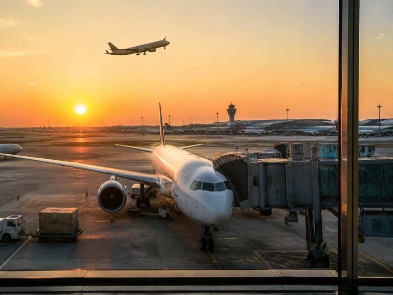 White passenger jet parked at a gate with a jet bridge attached during a sunset, other planes and a control tower visible on the tarmac in the distance.