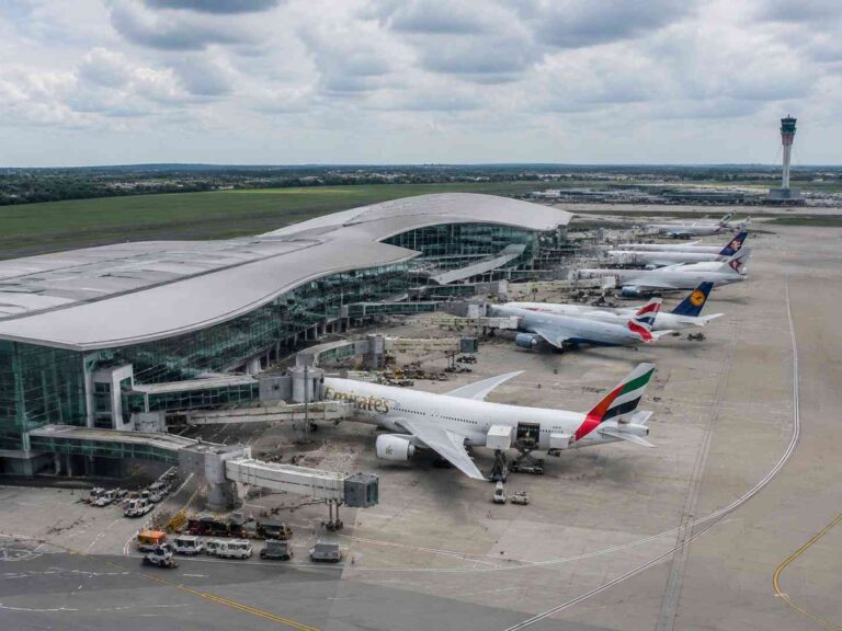 A wide view of a modern airport terminal with a row of parked international planes, including an Emirates jet at the gate, a Virgin/UK carrier and others lined up on the tarmac, with a control tower in the distance.