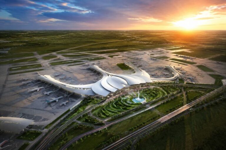 Aerial view of a modern airport terminal with curved white roofs, planes at gates, and a circular landscaped area with a fountain at sunset.