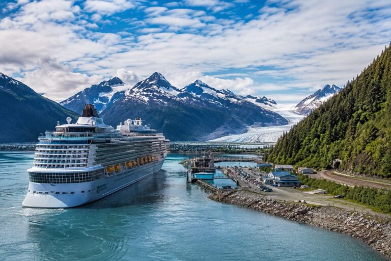 Cruise ship docked in a turquoise fjord with snow-covered mountains and a glacier in the distance.