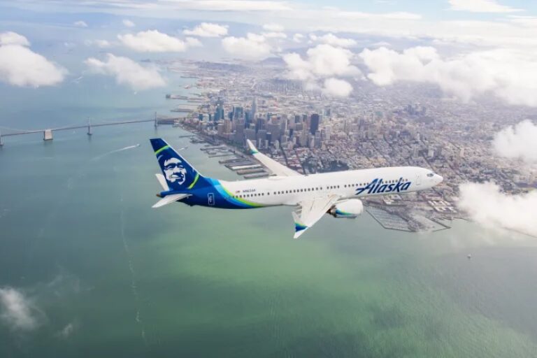 Alaska Airlines jet flying over a coastal city with a bay and bridge visible below, clouds in the sky.