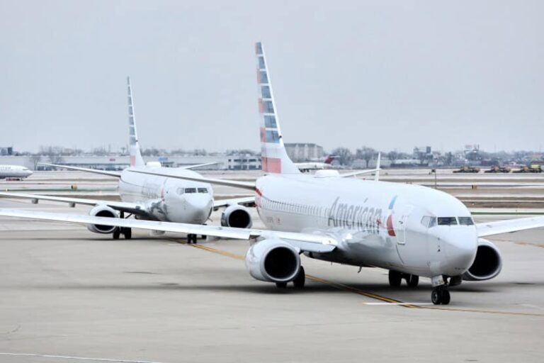 Two American Airlines airplanes on a taxiway at the airport, with distant terminal buildings visible in the background, engines idle or running slowly.
