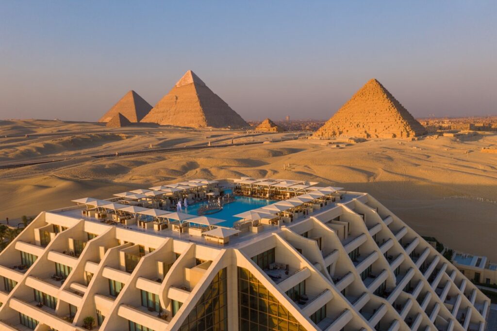 Modern white hotel with a rooftop pool and sun loungers, set in a desert with the pyramids of Giza in the background.