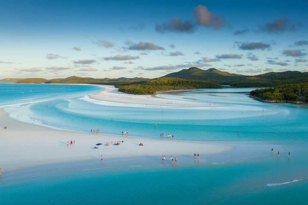 White-sand beach with turquoise lagoons and a forested peninsula; people stroll and sunbathe along the shore.