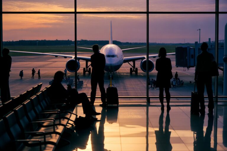 Silhouetted travelers waiting in a concourse as a plane sits at the gate at sunset.