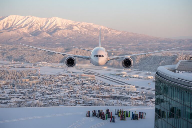 Airplane flying toward the camera over a snow-covered landscape with a snowy mountains backdrop and colorful ski poles in the foreground.