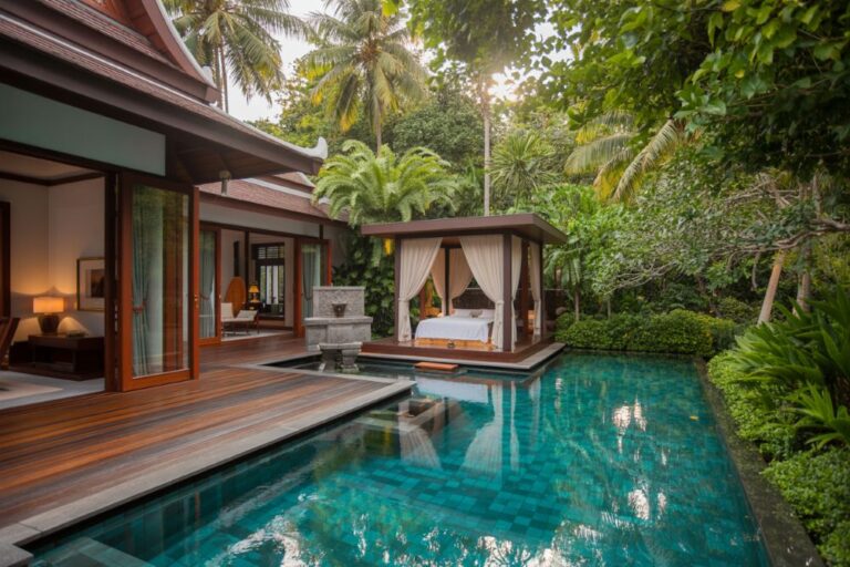 Luxury villa with a wooden deck, turquoise pool, and a canopy bed draped in white curtains among tropical foliage.