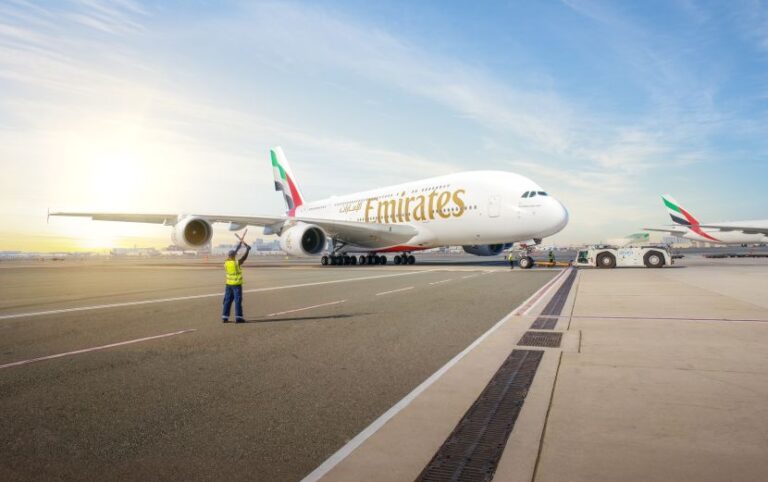 Ground crew member in reflective vest directs an Emirates A380 on the taxiway at sunrise with another plane in the background.