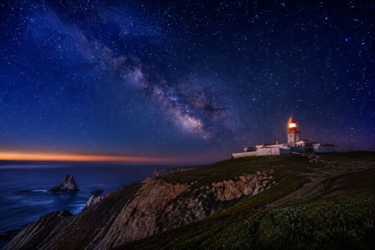 Star-filled night sky with the Milky Way over a coastal cliff; a lighthouse glows on a hill by the sea.
