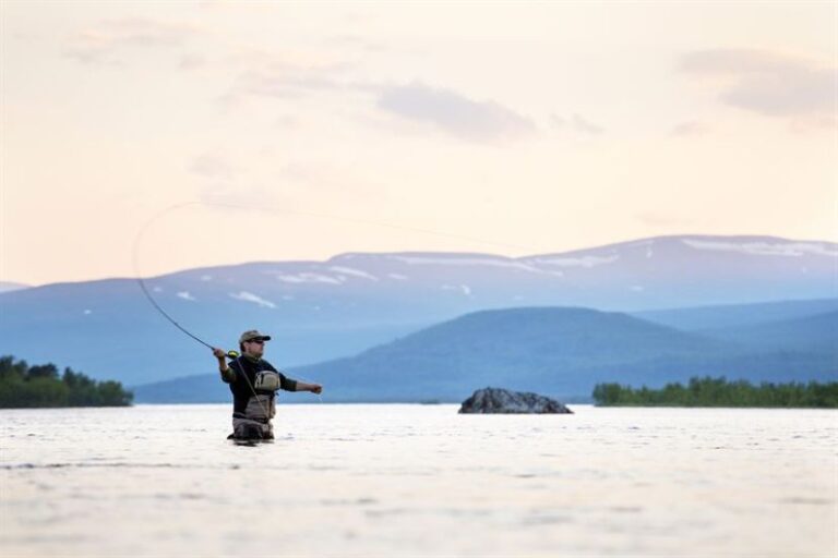 Man wearing waders fly-fishing in a calm lake, casting his line with mountains in the background at sunset.