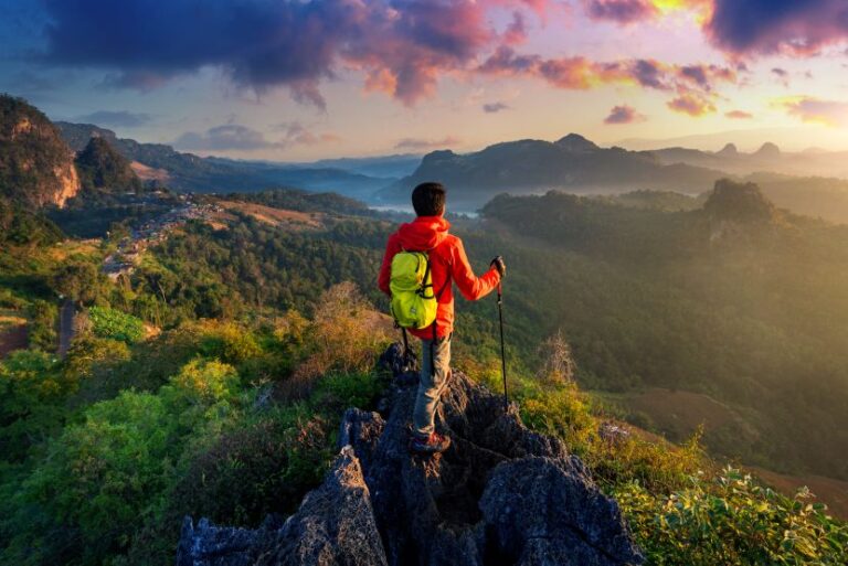 Hiker in a red jacket with a yellow backpack stands on jagged rocks, gazing over a sunlit valley at sunset.