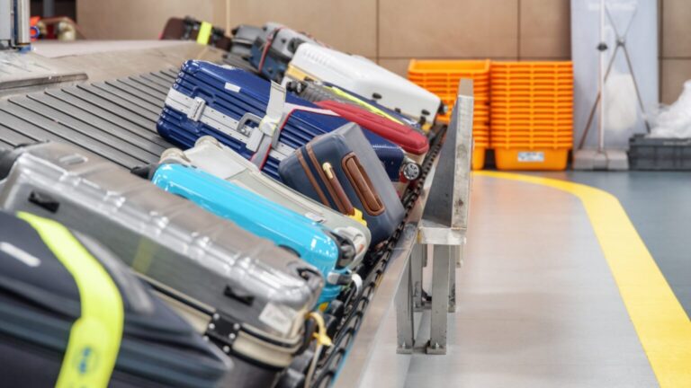 Baggage on a conveyor at an airport: colorful suitcases arranged on a metal belt, with orange storage bins in the background.