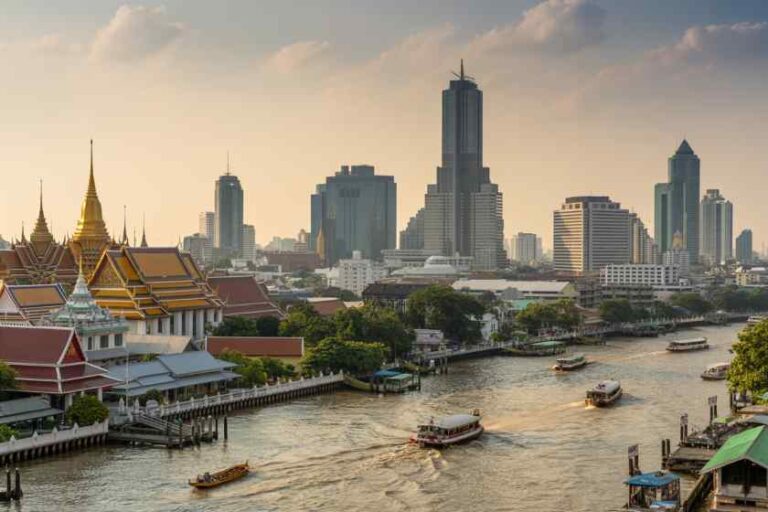Sunset view of Bangkok skyline with traditional temples along the Chao Phraya River and boats on the water.