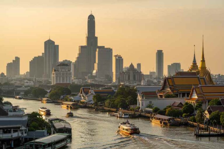 Bangkok skyline at sunset with boats on the Chao Phraya River and the Grand Palace complex on the riverbank.