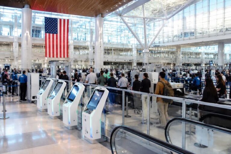 Busy airport check-in area with a row of self-service kiosks and travelers queuing under a large American flag.