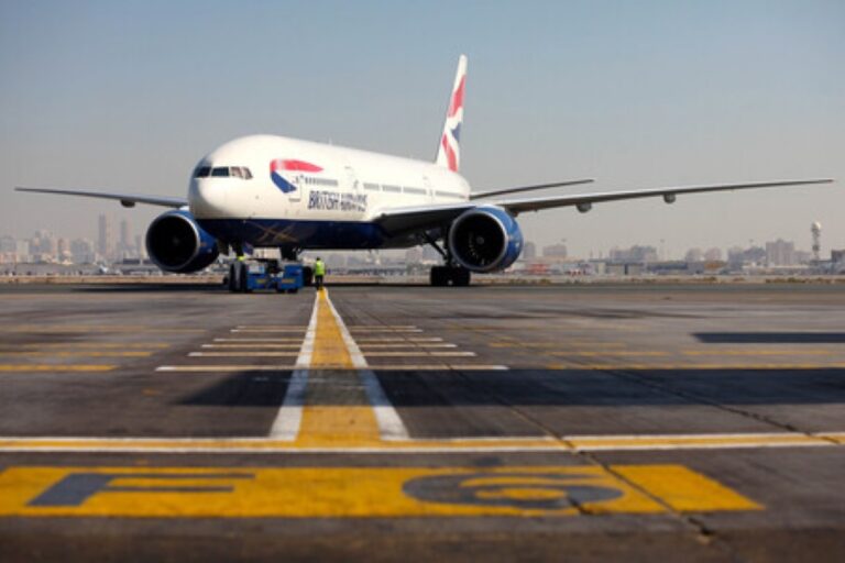 British Airways jet taxiing on a runway with ground crew nearby and a city skyline in the distance.