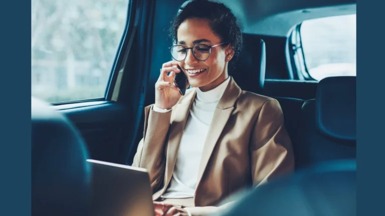 Woman in a tan blazer and glasses speaks on a cellphone while working on a laptop in the backseat of a car.