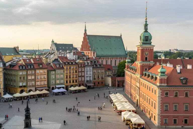 Historic European town square with colorful townhouses, outdoor cafes, and a large red-brick building with a green clock tower.