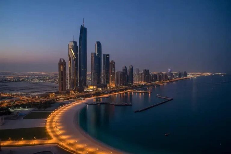 Evening view of a coastal city skyline with tall glass skyscrapers along a curved, illuminated marina promenade by the water.