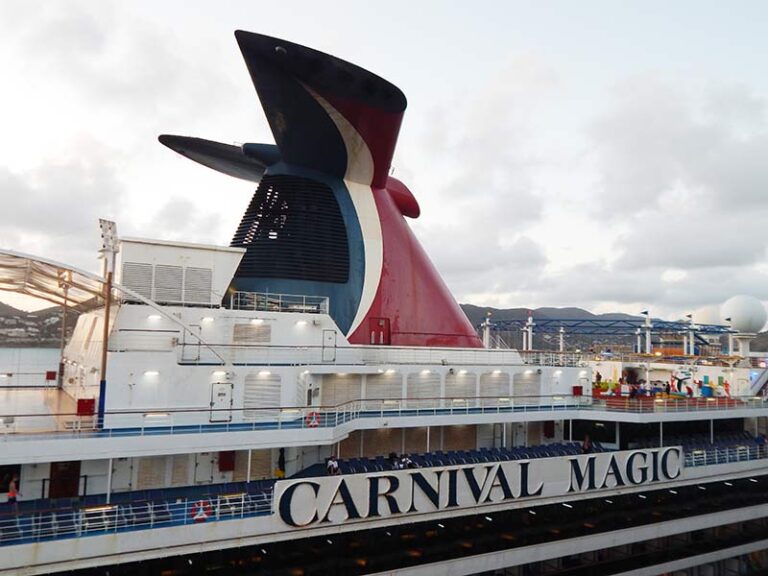 Side view of Carnival Magic cruise ship with a large colorful funnel and multiple decks steadied by railings and windows.