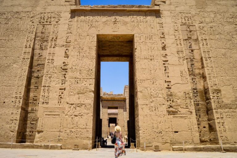 Tourist wearing a hat walks through a doorway of a sandstone Egyptian temple covered in hieroglyphs.