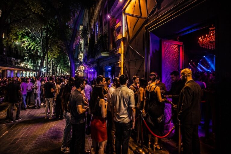 People queue outside a nightclub at night, with purple lighting and a chandelier visible inside the venue.