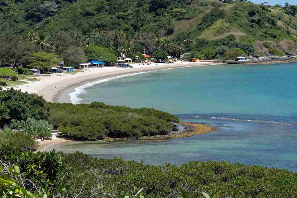 Tropical bay with a curved sandy beach, turquoise water, and palm-covered hills; small colorful huts line the shore.