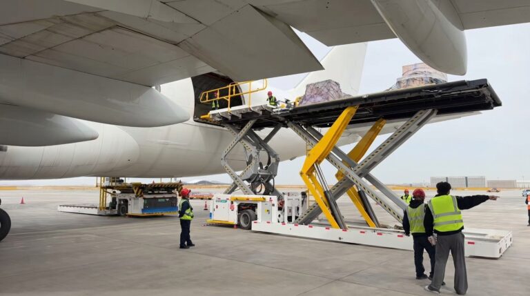 Ground crew in high-visibility vests load cargo from a yellow scissor lift into a large aircraft's open cargo door at the airport hall.igned