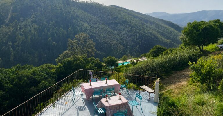 Outdoor dining terrace with pink tablecloths and turquoise chairs, overlooking a valley of pine-covered hills.