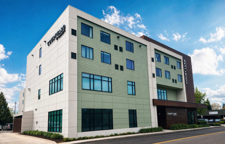 Exterior of a Courtyard by Marriott hotel building with white and green panels under a blue sky and scattered clouds, facing a street.