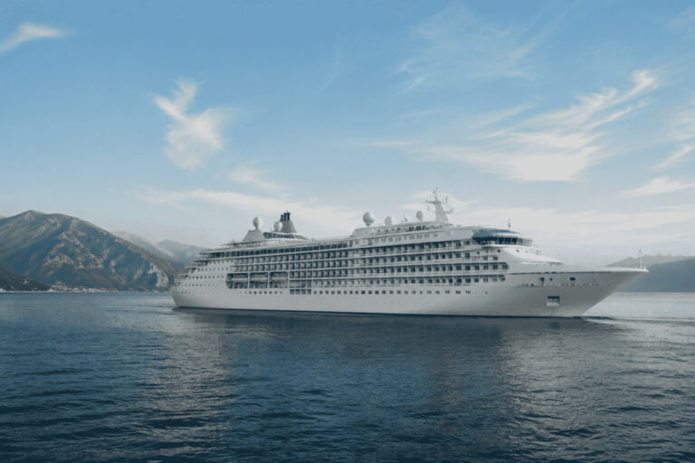 Large white cruise ship cruising on calm blue water with mountainous coastline in the background under a clear sky.