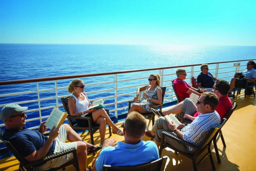 Group of passengers seated in outdoor deck chairs on a cruise ship, reading books as the ocean glitters nearby on a sunny day.