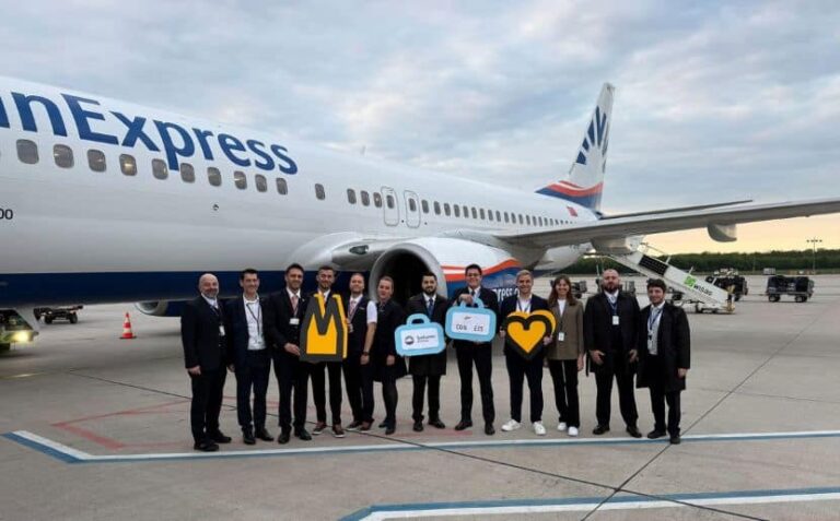 Group of airline staff posing on the tarmac in front of a parked plane, holding decorative signs like a crown, speech bubble, and heart icons.