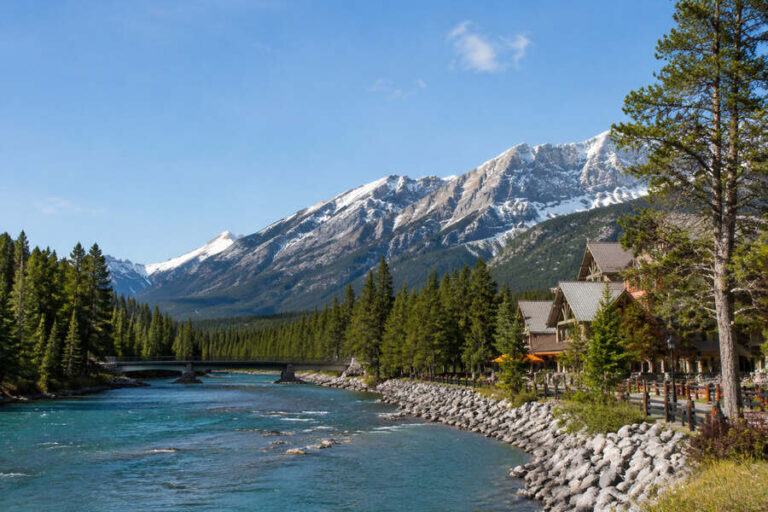 Snow-capped mountains overlook a turquoise river lined with pine trees and lodge-style buildings by the shore.