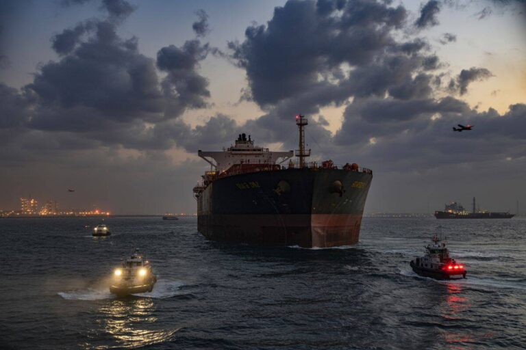 Large bulk carrier being guided by multiple tugboats across choppy sea at dusk, with industrial lights on the horizon and a plane in the sky.