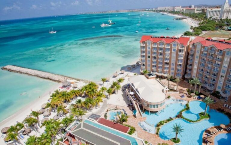 Aerial view of a beachfront resort with turquoise sea, palm trees, and a multi-story hotel surrounding a wavy swimming pool area.