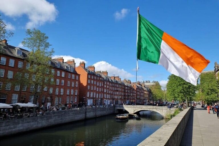 Ireland flag fluttering over a canal lined with red brick townhouses and a stone bridge under a blue sky.