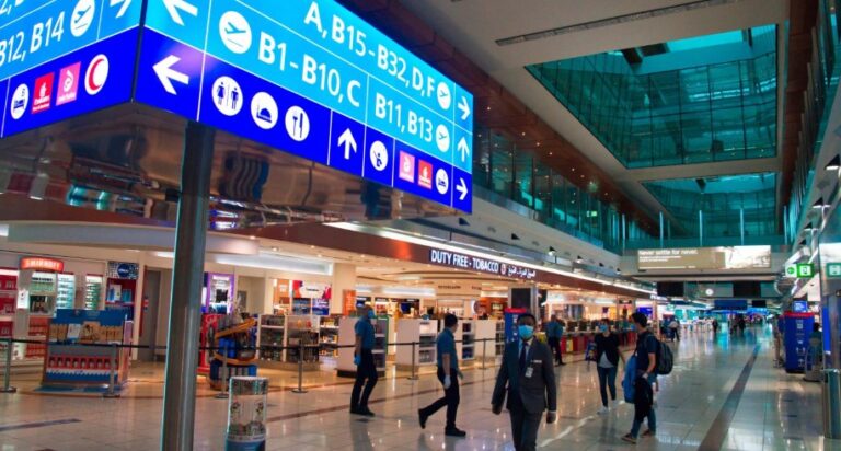 Busy airport terminal with bright blue overhead signs directing gates and services; duty-free shop visible in the background